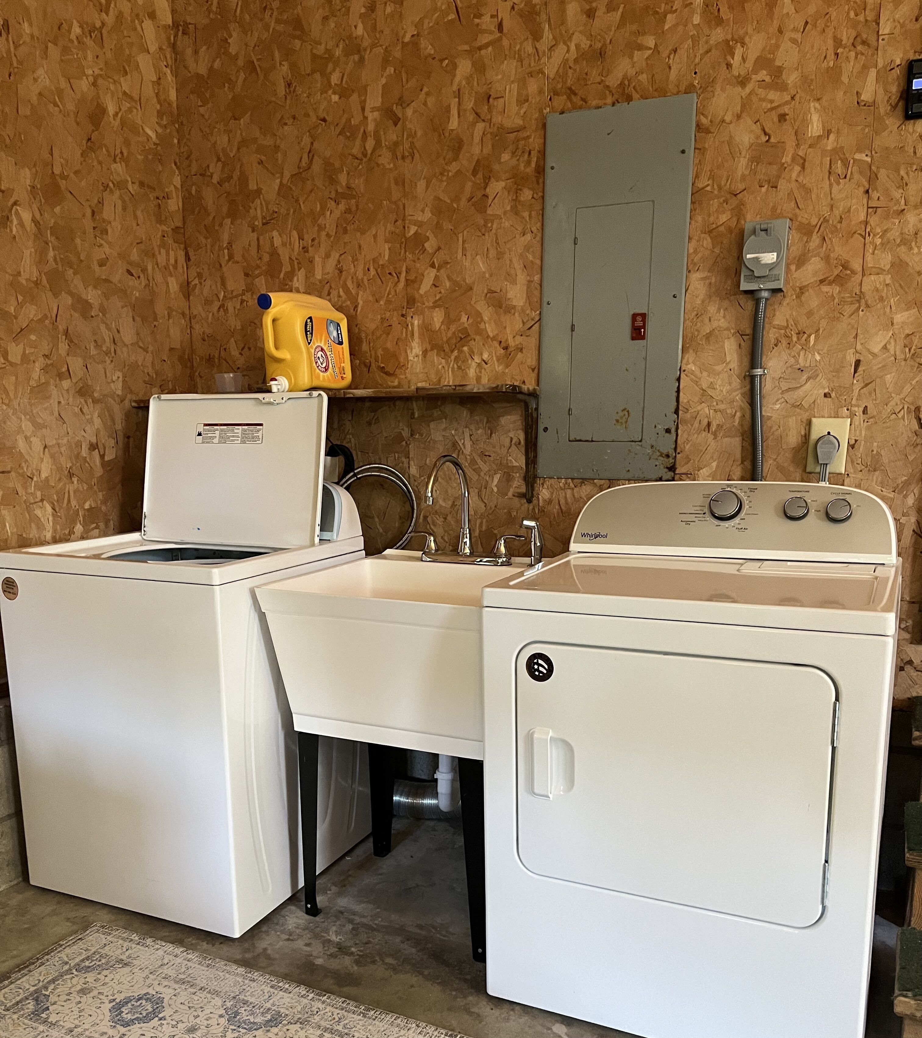 Une buanderie équipée de deux machines à laver et de deux sèche-linge Whirlpool blancs, d’un mur en bois, d’un compteur et d’une bouteille de lessive jaune posée sur le sèche-linge.