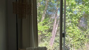 Une pièce dotée d’un plancher en bois, d’un lampadaire en osier, d’une chaise recouverte d’une couverture blanche et d’une porte-fenêtre coulissante donnant sur une terrasse extérieure bordée d’arbres.