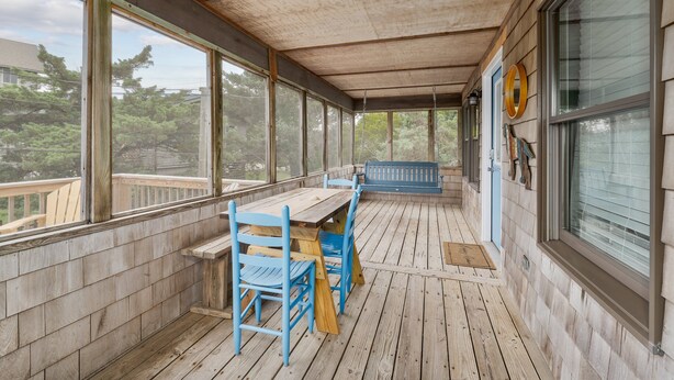 A covered porch with a wooden table and chairs, large windows, and a view of trees.