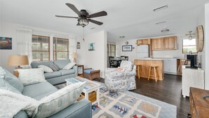 A spacious living area with a sofa, a patterned armchair, a coffee table, and a rug. A kitchen with wooden cabinets and a dining area with a table and chairs are visible in the background.