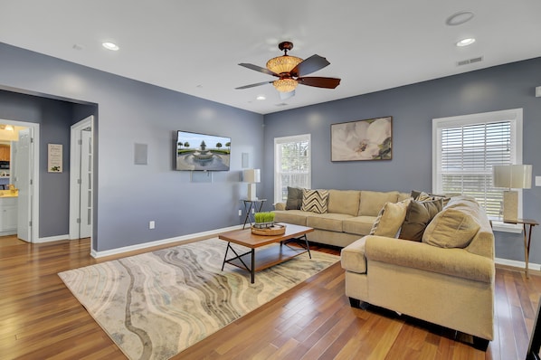 A living room with a beige sofa, a wooden coffee table, a ceiling fan, and a large window with blinds.