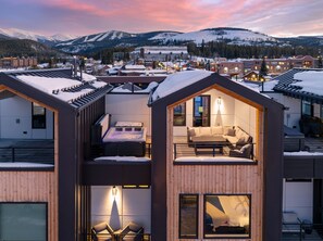 Une cabane moderne à plusieurs niveaux, dotée d’un bain à remous, d’un coin salon confortable et d’un balcon offrant une vue sur une ville de montagne enneigée.