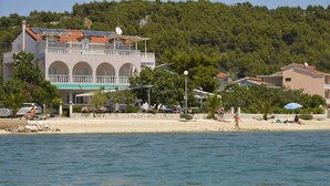 Ein Küstengebäude mit Balkon, ein Strand im Vordergrund und ein bewaldeter Hügel im Hintergrund.