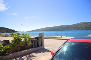 A parking area with a red car, a concrete barrier, and a view of a lake and hills. - 2-bed Apartment in Poljica Straight on Beach (Marina)