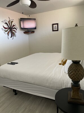 Uma cama bem arrumada com um edredom branco, uma televisão na parede, uma obra de arte decorativa e um abajur em uma mesa de cabeceira.