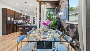 A modern kitchen with dark wood cabinets, a central island, and a dining area with a glass table and blue chairs. - Bloom House ● Luxe Floral Retreat with 12 Beds for Groups, Bachelorettes & ... (Houston)