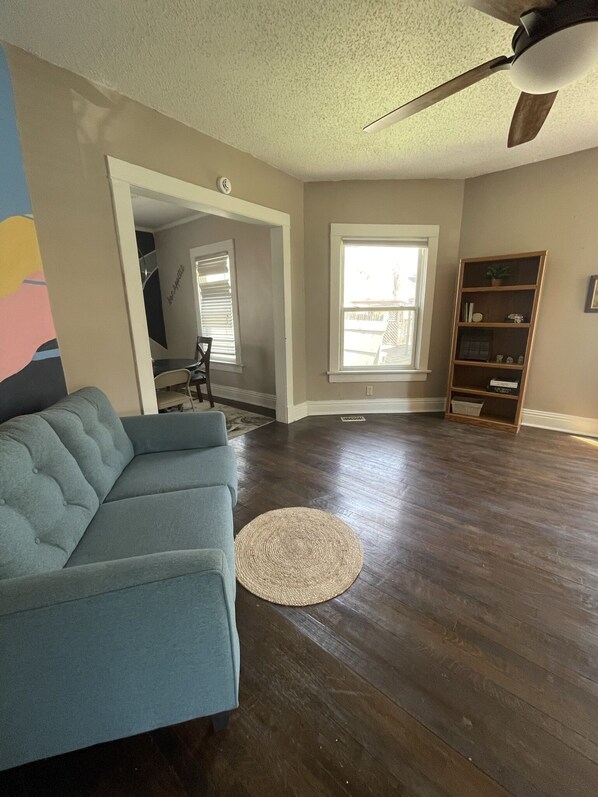 A living room with a blue sofa, a wooden floor, a ceiling fan, and a bookshelf.