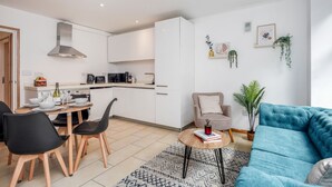 Une cuisine moderne avec des armoires blanches, une hotte de cuisinière en acier inoxydable et un coin repas avec une table dressée pour quatre personnes. Le salon est agrémenté d’un canapé bleu et d’une plante en pot.