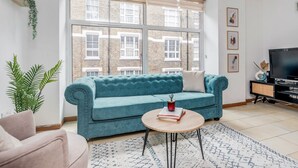 Un salon avec un canapé de couleur turquoise, une table basse ronde sur laquelle repose un livre, un meuble pour la télévision et une grande fenêtre donnant sur des immeubles en briques.
