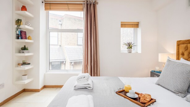 Une chambre d’hôtel bien rangée, avec un lit, une table de chevet dotée d’une lampe, une petite fenêtre avec une plante et une étagère contenant des livres et des objets décoratifs.