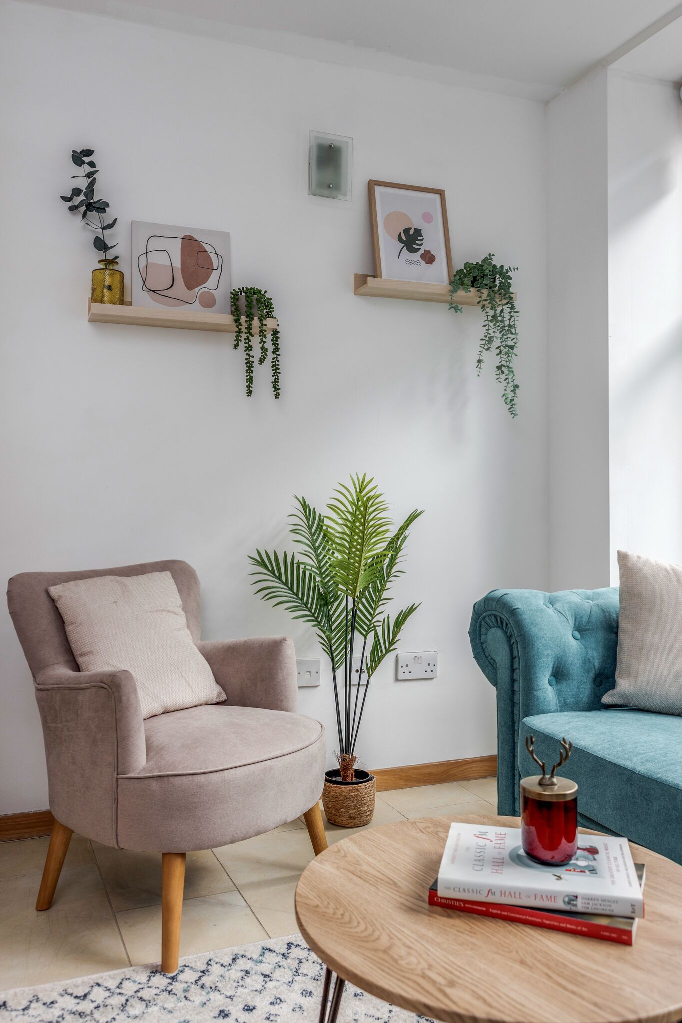 En stue med en beige lænestol, en blå sofa, et rundt træbord med bøger og en rød vase, samt vægmonterede hylder med planter og kunstværker.