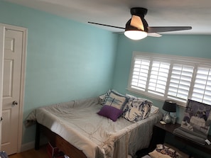 A bedroom with a bed, a ceiling fan, a window with blinds, and a bedside table with a lamp.