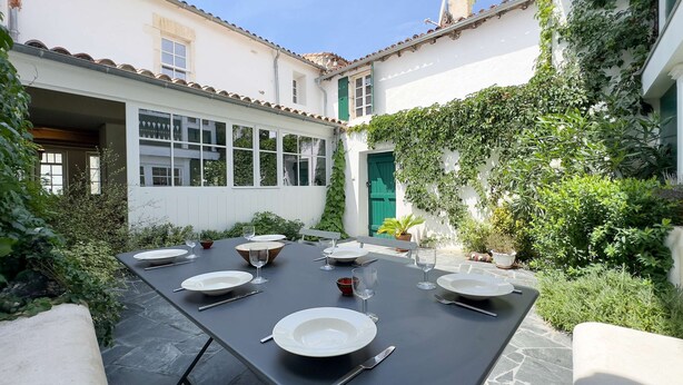 Mesa de comedor dispuesta para seis personas, con platos, vasos y cubiertos, rodeada de exuberante follaje y un edificio blanco con puerta verde.