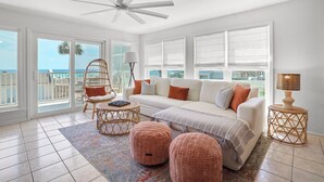 Un salon spacieux avec un canapé blanc, des fauteuils en osier, un ventilateur de plafond et de grandes fenêtres donnant sur la plage.