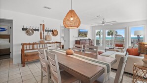 Un vaste espace de vie comprenant une table à manger en bois, des canapés blancs, un luminaire suspendu et une vue sur l’océan.