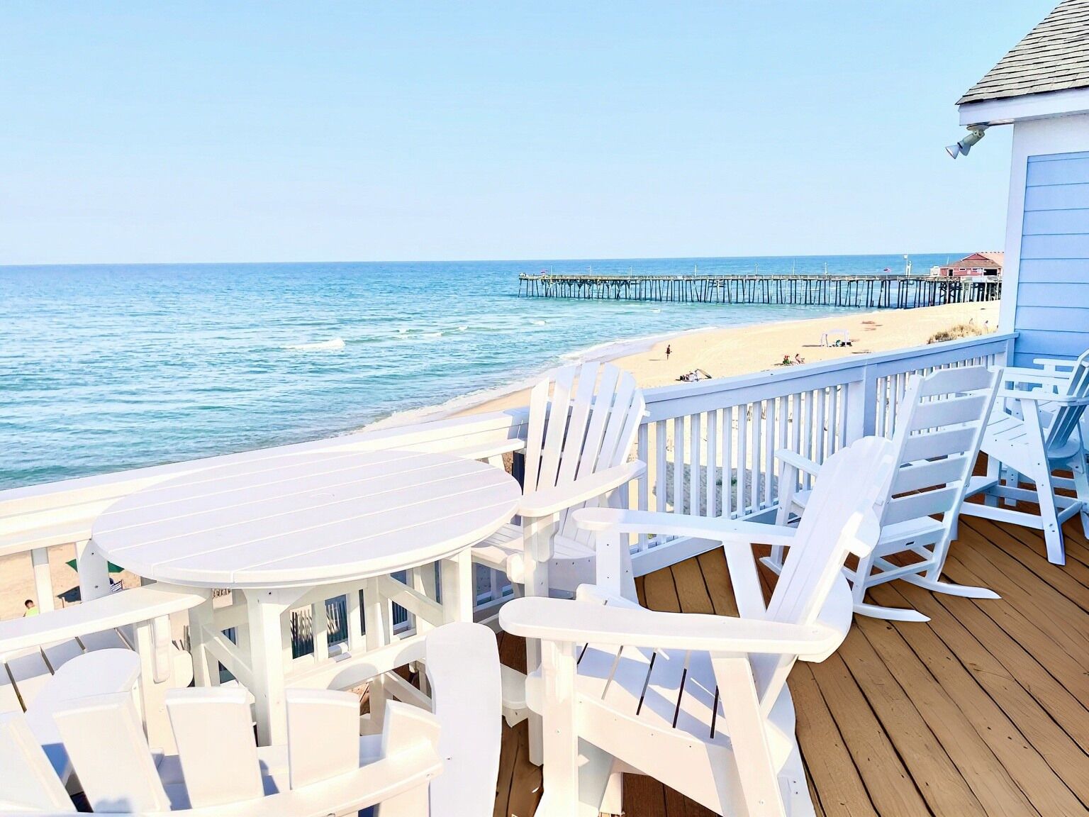 Een houten terras met witte stoelen en tafels met uitzicht op een strand en pier.