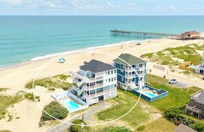 A beachfront property with a pool, multiple balconies, and a pier in the background. - Oceanfront Luxury Panoramic 5BR, Pool, Hot Tub, Elevator, Wave House OBX (Rodanthe)