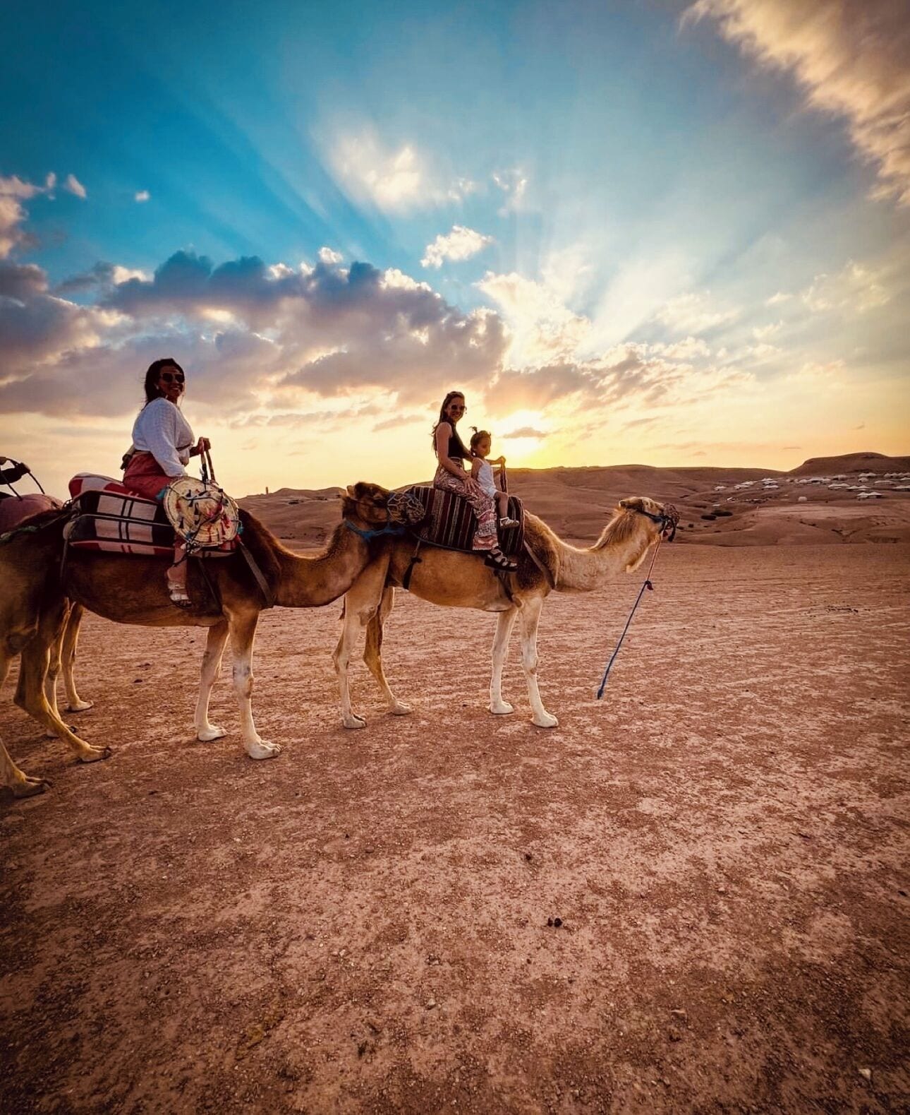 Dos camellos con sillas de montar y jinetes en un paisaje desértico durante el atardecer.
