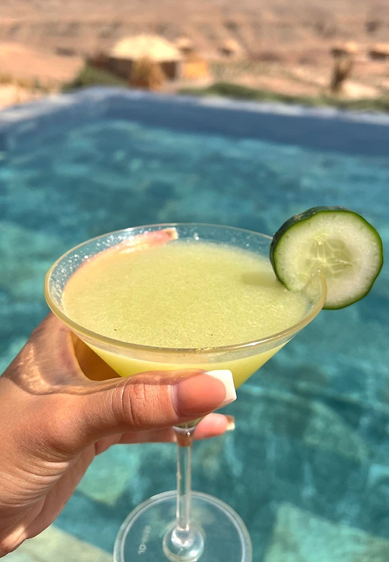Una mano sostiene una copa de cóctel con una rodaja de pepino, frente a una piscina.