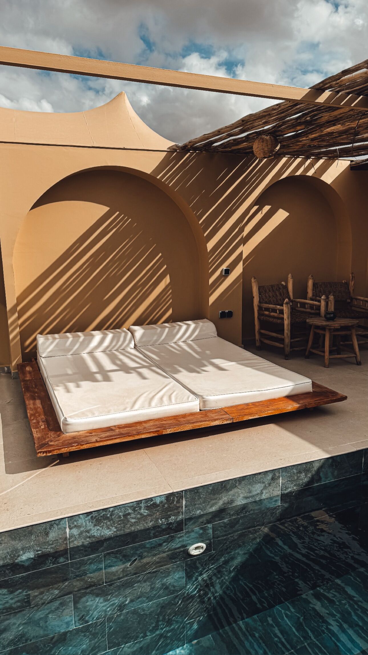 A single bed with a wooden frame and white bedding, positioned on a balcony with a view of a swimming pool.