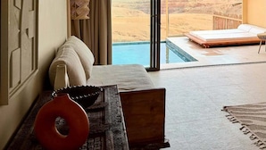 Ein Zimmer mit großem Fenster mit Blick auf eine Wüstenlandschaft, ein Bett, ein kleiner Tisch mit roter Vase und ein gemustertes Teppich.