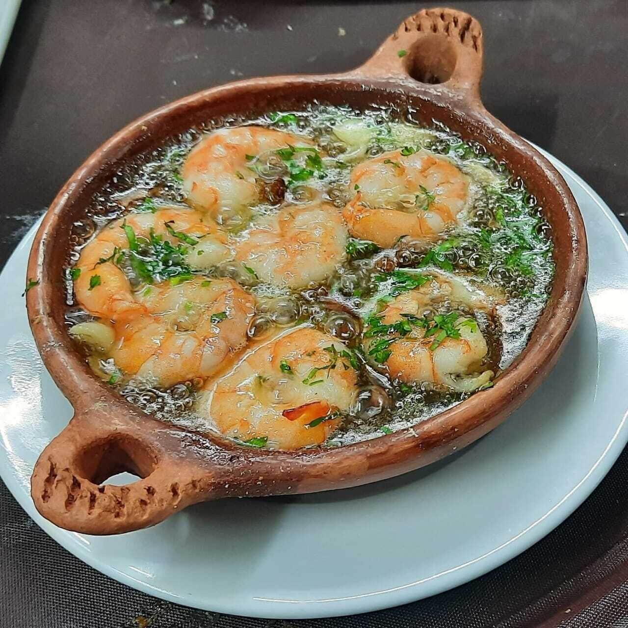 A clay pot with shrimp cooking in oil, garnished with herbs.