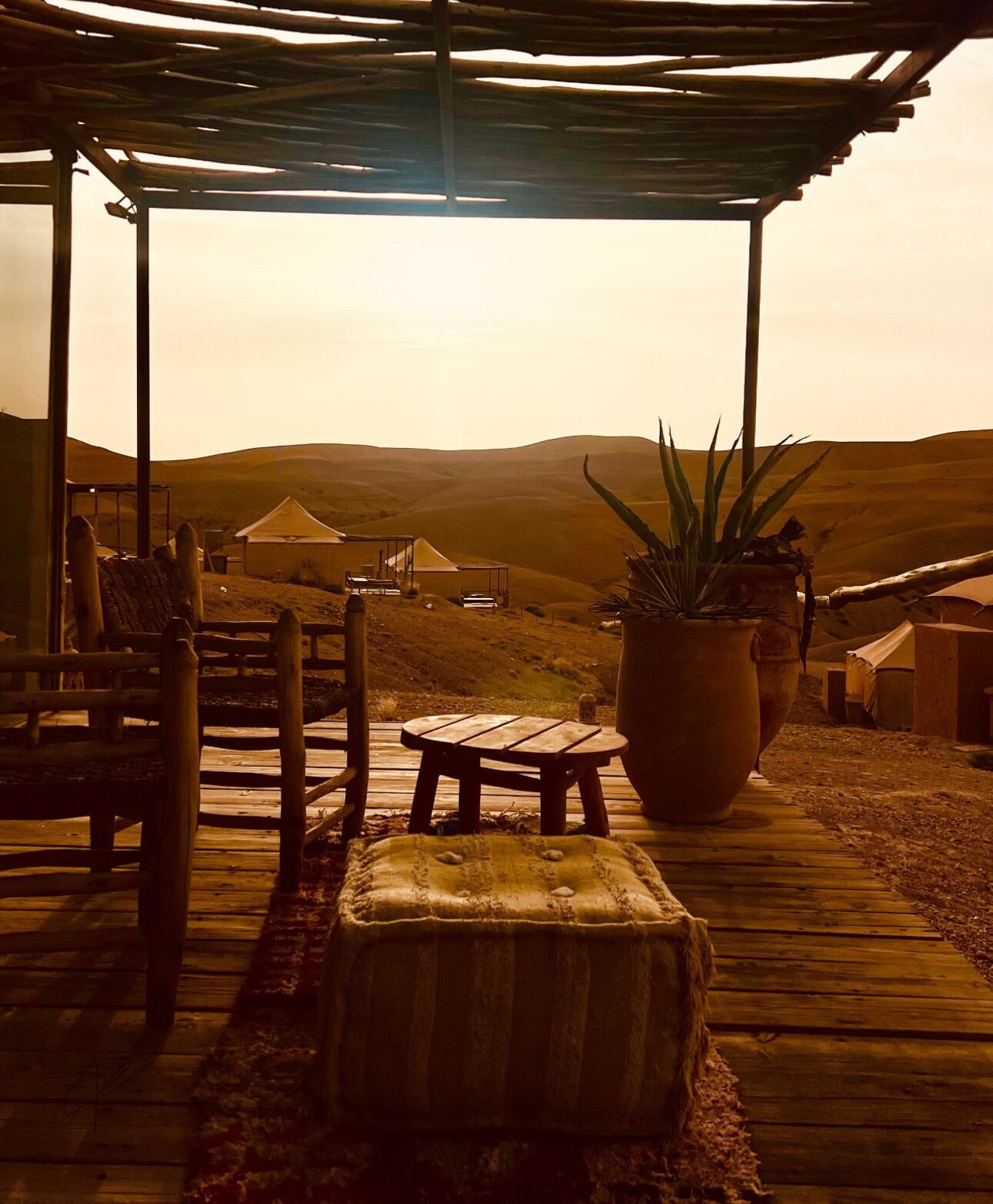A wooden deck with a covered seating area, a large potted plant, and a view of tents in the background.