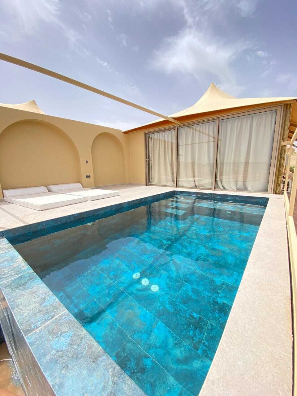 A rectangular swimming pool with a clear blue tint, surrounded by beige walls and a covered area with white curtains.