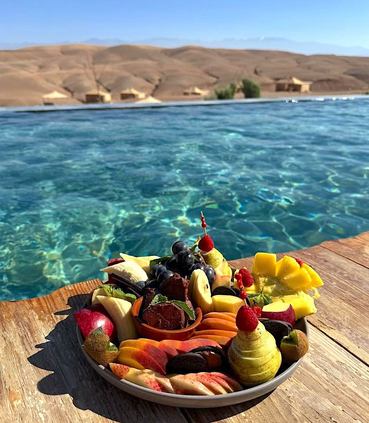 A fruit platter on a wooden table by a pool with clear water and a mountainous backdrop.
