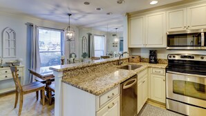 A kitchen with white cabinets, a granite countertop, stainless steel appliances, and a dining area with a wooden table and chairs. - 938 Cutter Court at Sea Pines Resort (Hilton Head Island)