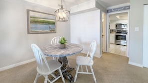 A dining area with a round table, white chairs, a painting of a landscape, and a kitchen visible through an open door. - 955 Cutter Court at The Sea Pines Resort (Hilton Head Island)