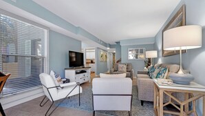 A modern living room with a sofa, armchair, and a coffee table. There is a TV and a dining area with a lamp.