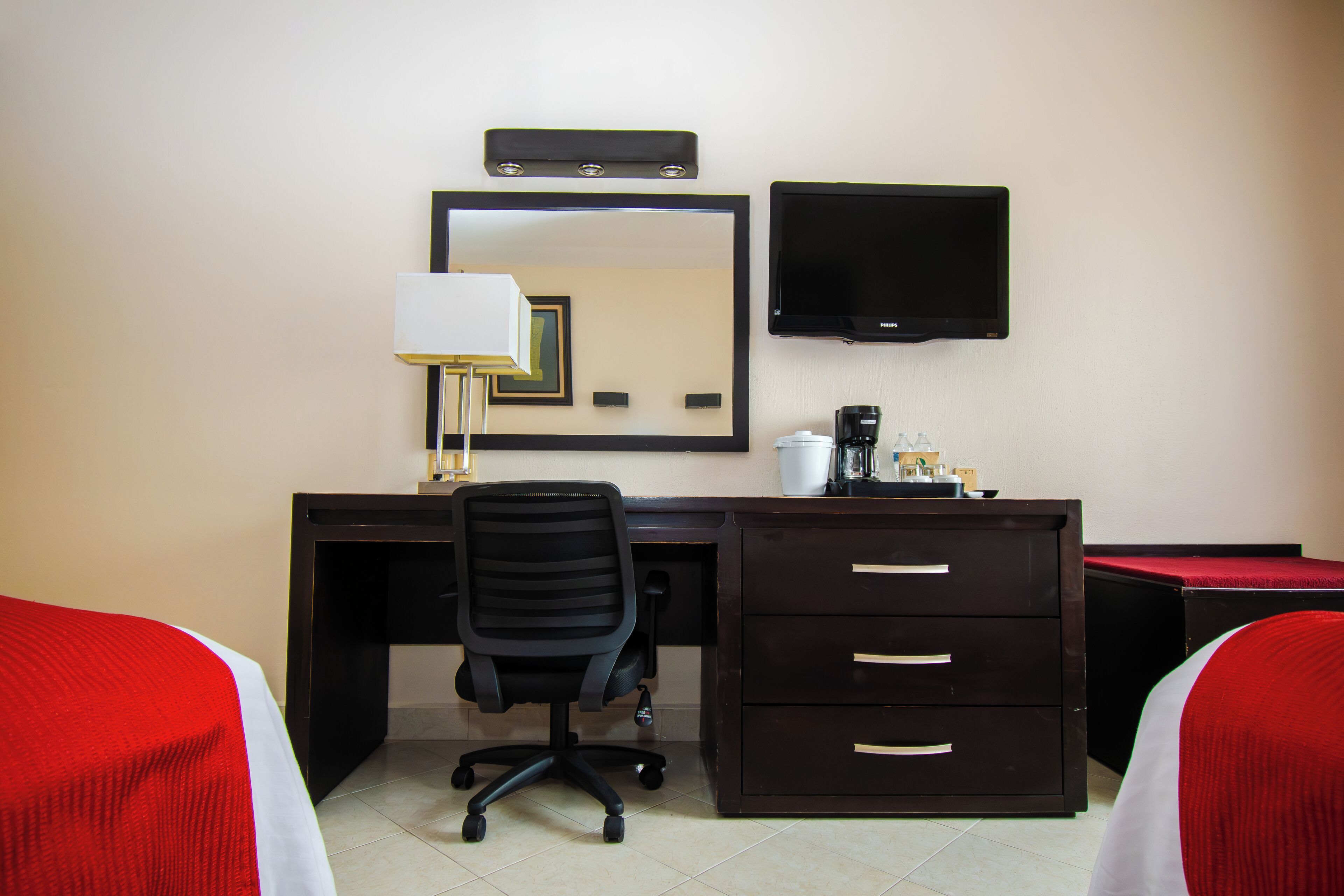 Habitación de hotel con escritorio, una silla, televisor, espejo y dos camas con ropa de cama roja y blanca.