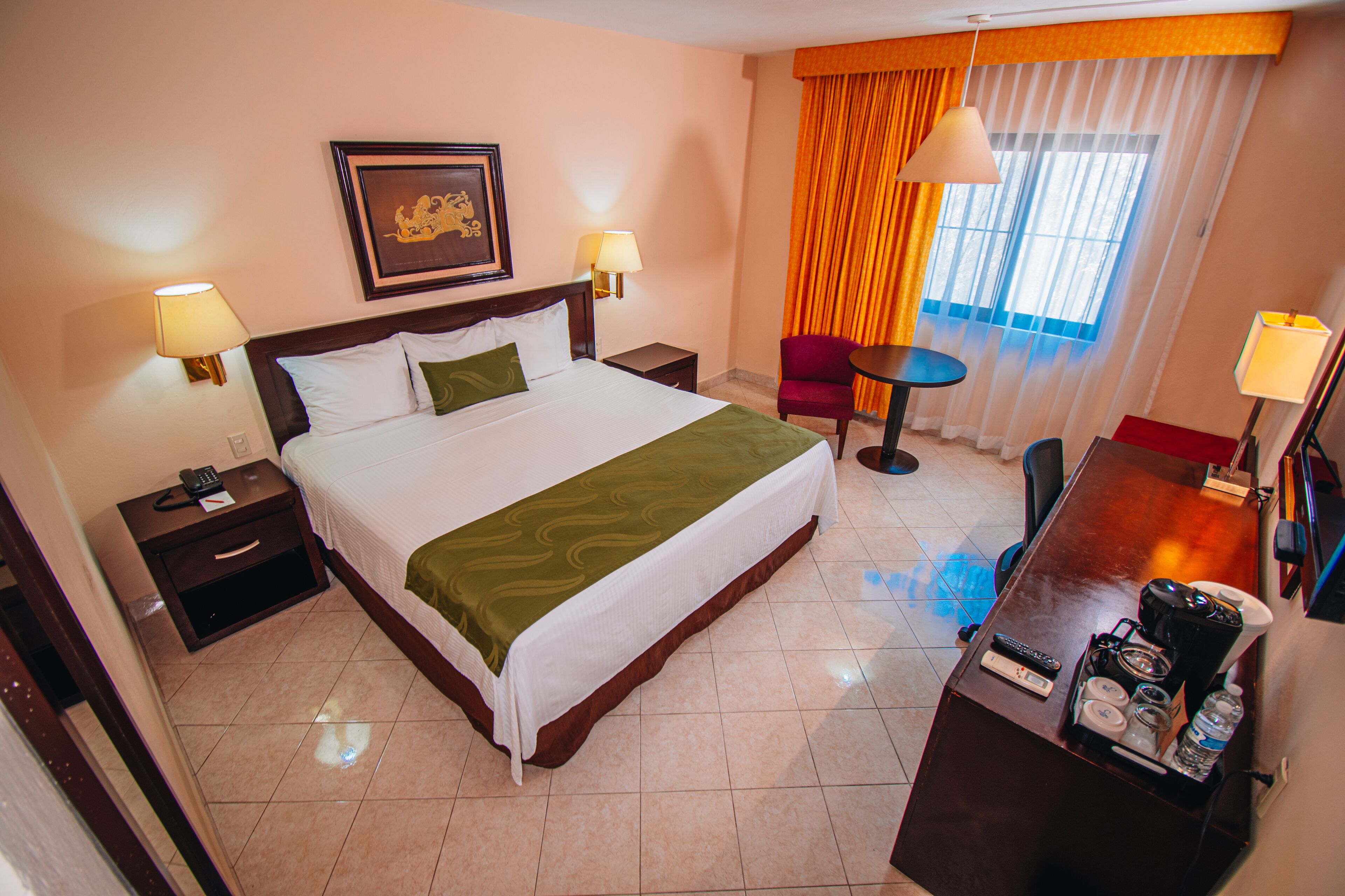 Una habitación de hotel con una cama grande, dos mesitas de noche, un escritorio con silla, una mesita pequeña y una ventana con cortinas.