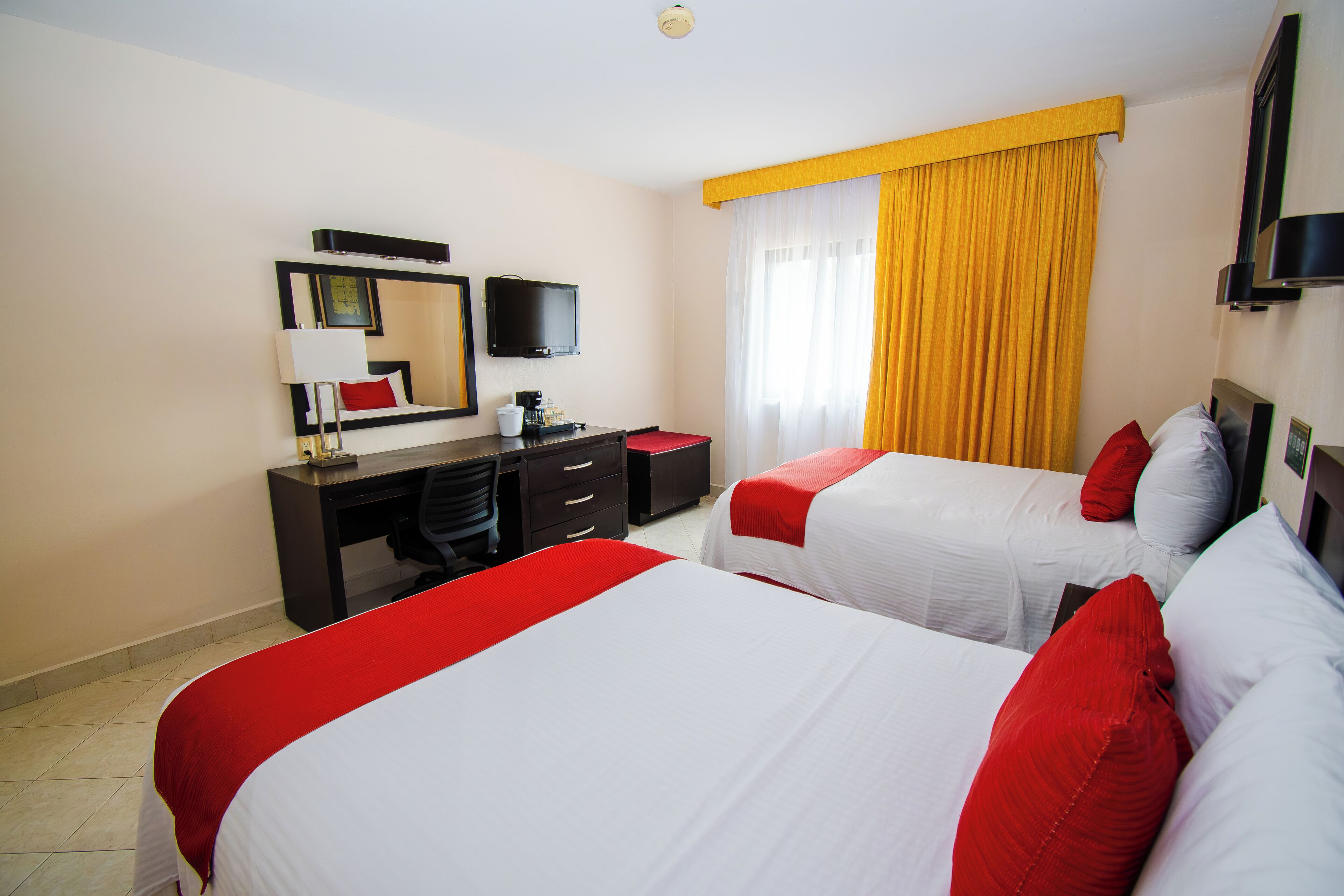 A hotel room with two beds, a desk, a chair, a TV, and a window with yellow curtains.
