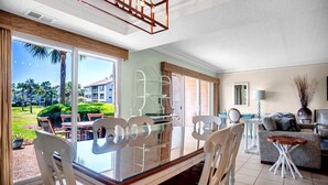 Un vaste espace de vie comprenant une table à manger en verre, des chaises blanches, un canapé gris et une vue sur un jardin luxuriant et un bâtiment.