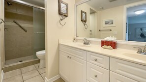 Une salle de bain avec douche, toilettes, un meuble-lavabo avec tiroirs, un grand miroir et des tableaux encadrés accrochés au mur.