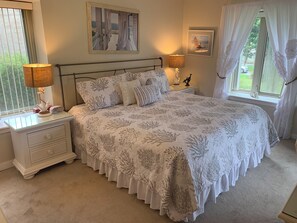 Une chambre à coucher avec un lit, des tables de chevet, une lampe, une fenêtre avec des rideaux et un tableau encadré au mur.