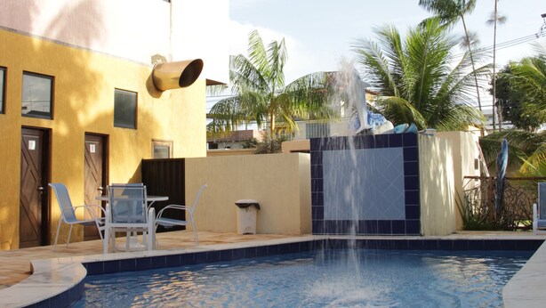 A swimming pool with a water fountain, surrounded by palm trees and a building with a tiled roof. - Pousada Vila do Porto (Ipojuca)