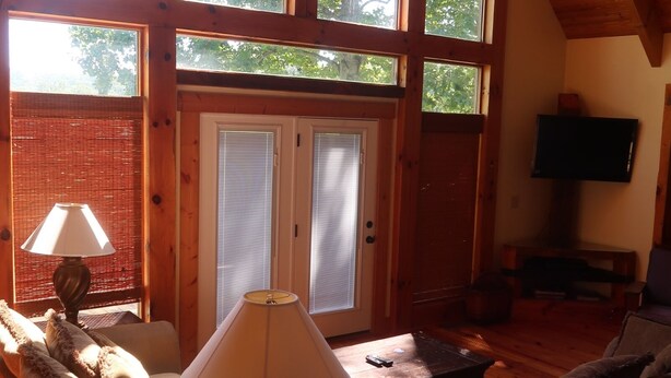 Ein gemütliches Wohnzimmer mit Holzboden, einer Couch, einem Couchtisch und einer Lampe. Große Fenster lassen natürliches Licht und Blick auf Bäume herein.