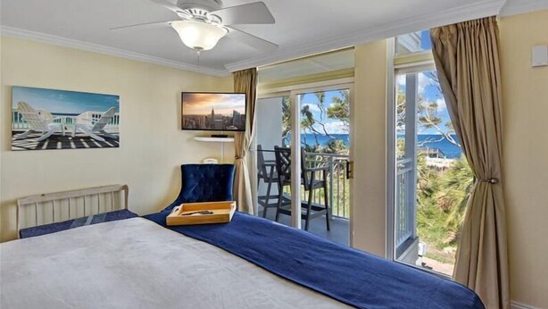A hotel room with a bed, a ceiling fan, a TV, and a balcony with a view of the ocean.
