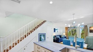 A modern kitchen with a marble countertop and a stainless steel sink, a staircase with white railings, and a living area with blue furniture and a view of the outdoors.