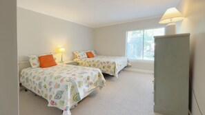 Un dormitorio con dos camas, cada una con una colcha colorida y una almohada naranja. Hay una ventana con persianas, una lámpara en la mesita de noche y un armario.