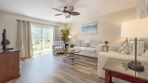 A spacious living room with a large window, a ceiling fan, a sofa, a coffee table, and a TV on a stand. - 2270 Heritage at The Sea Pines Resort (Hilton Head Island)