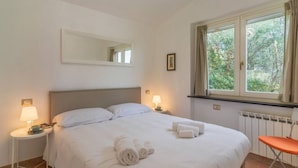 Una cama bien hecha con toallas enrolladas, una mesita de noche con lámpara, una ventana con cortinas y un radiador.