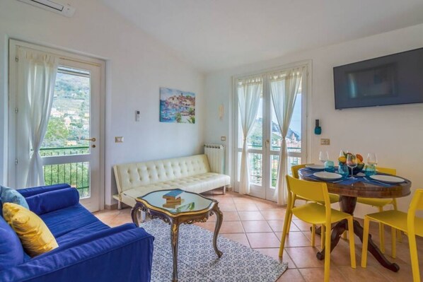 Un soggiorno con divano, tavolo, sedie gialle, televisione e balcone con tende.