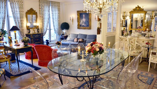 A spacious living room with a glass-top dining table, clear chairs, a red sofa, and a chandelier.