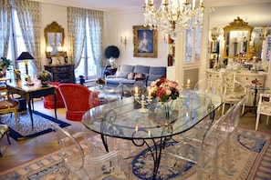 Uma sala de estar ampla, com uma mesa de jantar de vidro, cadeiras transparentes, um sofá vermelho e um lustre.