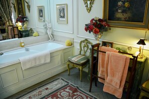 Une salle de bain avec une baignoire, une chaise, une coiffeuse avec un miroir et un vase de fleurs.