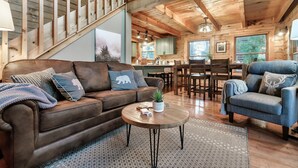 A cozy cabin interior with a wooden staircase, a brown leather sofa, a blue armchair, and a dining area with wooden furniture. - Stunning Sunset Views! Hot Tub, 2 Fire Pits, Game Loft, Dogs Welcome, Pool Open! (Sevierville)